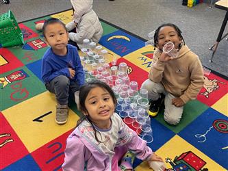 Three kindergarten students smile at the camera while they crouch on a colorful classroom carpet. They are sitting around a stack of clear plastic cups.