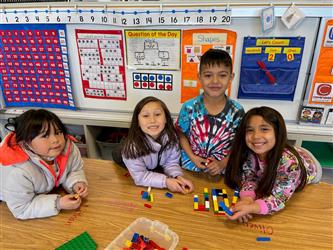 Four kindergarten students sit at a table and smile at the camera. One is sitting off to the side. The other three are close to each other and are holding an item they made out of legoes.