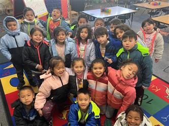 Four kindergarten students sit at a table and smile at the camera. One is sitting off to the side. The other three are close to each other and are holding an item they made out of legoes.