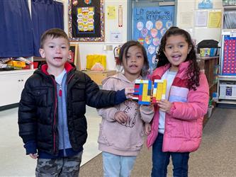four female students stand and smile at the camera. They are holding items they made out of legos. A classroom is in the background.