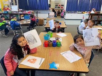 four female students stand and smile at the camera. They are holding items they made out of legos. A classroom is in the background.