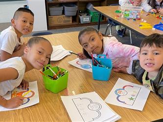 A group of kindergarten students pose behind a tower they made out of blocks. They are smiling at the camera and a classroom is in the background.