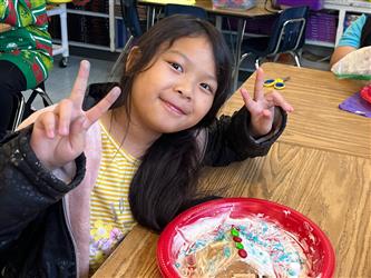 a student smiling at the camera and holdng up peace signs on both hands while sitting at a desk with a gingerbread house on a plate