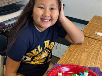 a student smiling at the camera while sitting at a desk with a gingerbread house on a plate