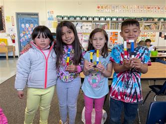 Four kindergarten students sit at a table and smile at the camera. One is sitting off to the side. The other three are close to each other and are holding an item they made out of legoes.